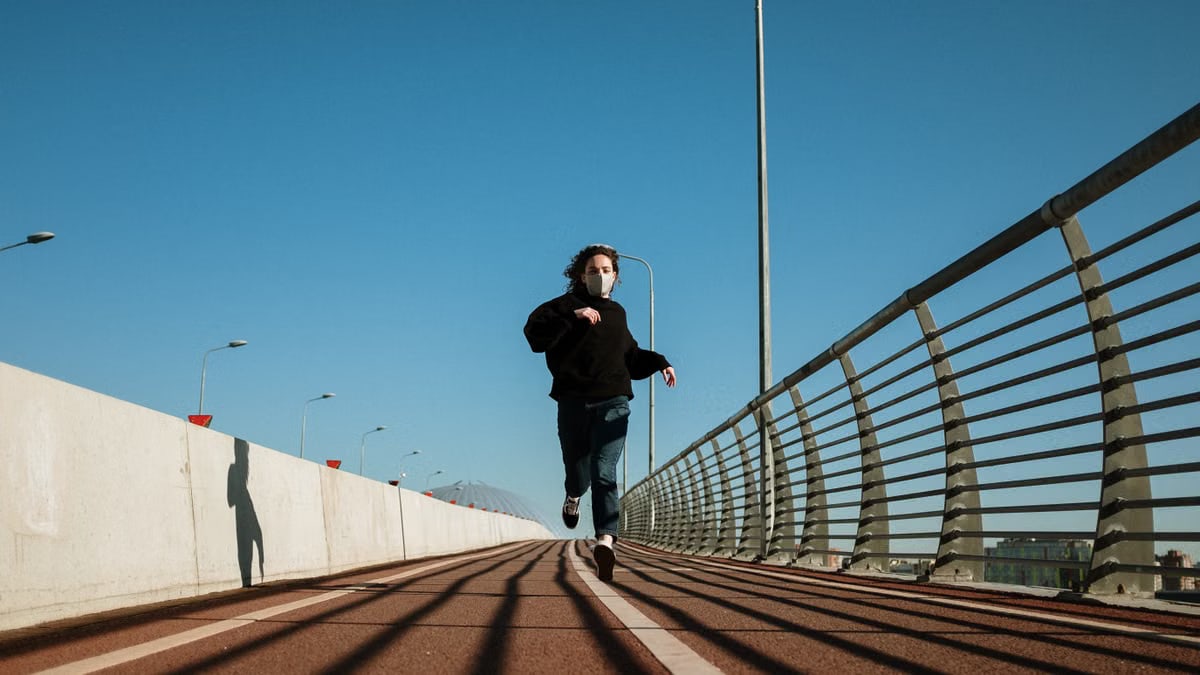 A person wearing a face mask runs alone on an urban bridge pathway under a clear blue sky.